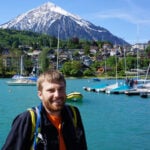 A man with a beard stands by a turquoise lake with sailboats and a small town in the background. A snow-capped mountain rises behind the town under a clear blue sky.