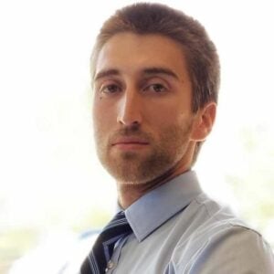 A man with short brown hair and a beard, wearing a light blue shirt and a dark blue tie, looks directly at the camera with a neutral expression. The background is blurred and bright.